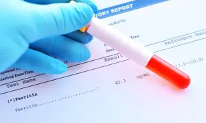 Ręka w niebieskiej rękawiczce trzyma probówkę z krwią nad raportem laboratoryjnym. Wynik ferrytyny 62.3 ng/mL mieści się w normie.