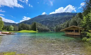 Ein idyllischer See mit türkisfarbenem Wasser, umgeben von grünen Hügeln und Wäldern. Ein Holzsteg mit Booten und ein kleines Holzhaus am Ufer laden zum Verweilen ein. Perfekt für einen blindsee trail.