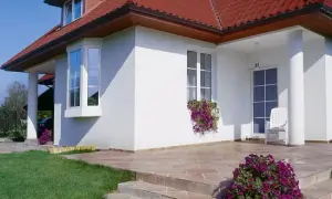 Elegancki dom z białym tynkiem, czerwonym dachem i charakterystycznym wykuszem. Przed wejściem kwitną fioletowe kwiaty.
