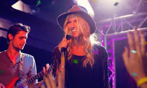Cantanti marchigiani sul palco, una donna con cappello canta al microfono, un chitarrista suona.