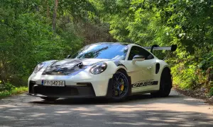 Biały Porsche 911 GT3 RS z czarnymi pasami na masce i bokach, zaparkowany na leśnej drodze. Jego cena jest wysoka, ale wrażenia z jazdy bezcenne.