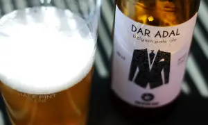 Bicchiere di birra dorata con schiuma bianca e bottiglia di Dar Adal belgian pale ale con etichetta a forma di giacca.