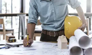 Ein Bauleiter, der als Generalunternehmer Baupläne prüft. Er hält einen gelben Helm und einen Stift.