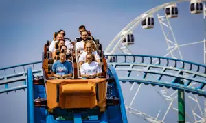 Ludzie na wagoniku rollercoastera przeżywają emocje podczas zjazdu. W tle widać koło widokowe.