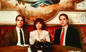 Jeremy Irons w filmach, tutaj z dwiema innymi osobami, siedzi przy stole w eleganckiej restauracji.