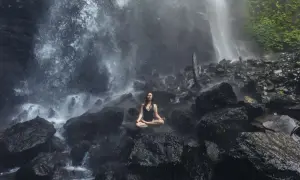 Mujer meditando en posición de loto sobre rocas, rodeada por la bruma de una cascada. Un momento de sanación pránica en la naturaleza.