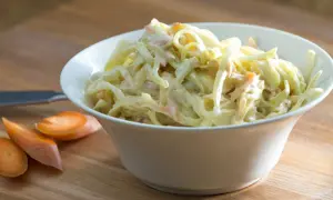 En skål med krämig, godaste coleslaw med strimlad vitkål och morötter. Bredvid ligger några morotsskivor.