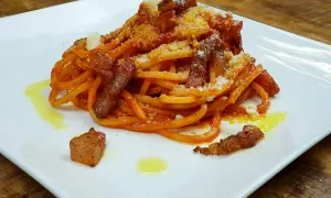 Bucatini con salsa amatriciana, cubos de guanciale crujiente y queso rallado, servido en plato blanco.
