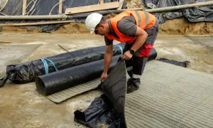 Budowlaniec rozkłada warstwę **betonitu** na placu budowy. Obok leżą zrolowane materiały izolacyjne.