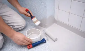 Person målar våtrumsmatta med pensel och roller. En hink med färg står bredvid.