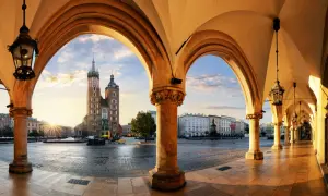 Krakau Geheimtipps: Blick durch Arkaden auf den Marktplatz mit Marienkirche im Sonnenaufgang. Ein malerischer Ort für Entdeckungen.