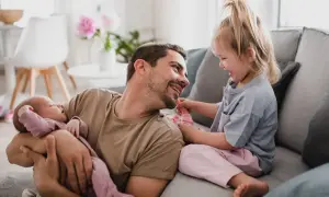 Szczęśliwy ojciec na urlopie tacierzyńskim bawi się z córką i tuli śpiące niemowlę.