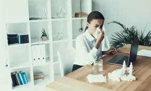 Kobieta z chusteczką przy nosie siedzi przy biurku z laptopem. Na stole leżą leki i chusteczki. To chyba jej ostatni dzień pracy, l4 na wypowiedzeniu.