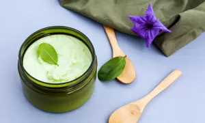 Un pot de gommage cuir chevelu maison, vert pâle, avec des cuillères en bois et une fleur violette.