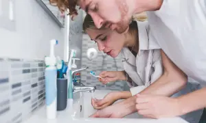 Para młoda nad umywalką. Ona płucze szczoteczkę, on pochyla się nad nią. Czy powinno się płukać zęby po umyciu?