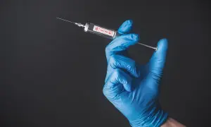 Ręka w niebieskiej rękawiczce trzyma strzykawkę z napisem VACCINE. To symbol szczepień, być może w kontekście Tunezji.