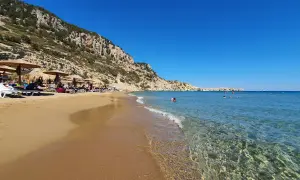 Słoneczna plaża Tsambika z piaskiem, leżakami i parasolami pod skalistym zboczem. Krystalicznie czysta woda zaprasza do kąpieli.