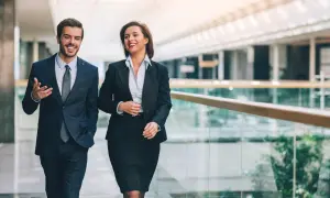 Ein Mann und eine Frau in formeller Geschäftskleidung gehen lächelnd durch ein modernes Bürogebäude.