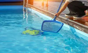 Person mit Kescher im Wasser, um den Pool sauber machen. Ein gelbes Spielzeug treibt im klaren Wasser.