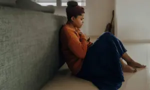 En ung kvinna sitter på golvet, lutad mot en soffa, med händerna över bröstet. Hon ser ut att vara i djup tanke, kanske under en gad utredning av sina känslor.