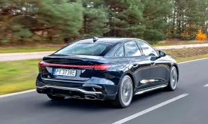 Ciemny Audi A4 B10 w wersji sedan pędzi po drodze, otoczony jesiennym lasem.