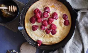 Puszysty omlet z malinami, posypany cukrem pudrem, podany w żeliwnej patelni. Obok miseczka z miodem i plastrem pszczelim.