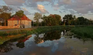 Dworek w Gdańsku Olszynce z czerwonym dachem odbija się w spokojnej wodzie kanału. Chmury na niebie malują zachód słońca.