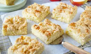 Saftiger Apfelkuchen vom Blech mit Streuseln, frisch gebacken und in Stücke geschnitten. Daneben liegen Äpfel.