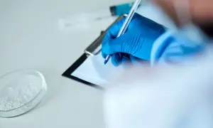 Ręka w niebieskiej rękawiczce pisze na kartce. Obok szalka Petriego z substancją i strzykawka. To może być test na narkotyki.