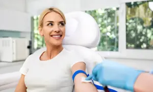 Kobieta w białej koszulce uśmiecha się, gdy pielęgniarka pobiera jej krew. To jeden ze sposobów, jak zbadać progesteron.