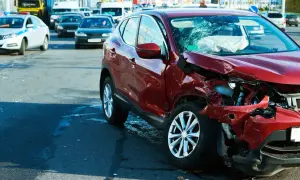 Wypadek drogowy. Czerwony samochód po kolizji, zniszczony przód i pęknięta szyba. Na drodze inne pojazdy, w tym radiowóz. Może to być sytuacja wymagająca **umowy na zastępstwo** kierowcy.