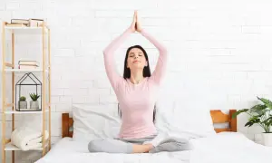 Mujer meditando en la cama, con las manos juntas sobre la cabeza, practicando dinámicas de relajación.