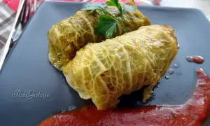 Dwa gołąbki z ryżem i mięsem wieprzowym, polane sosem pomidorowym, ozdobione natką pietruszki.