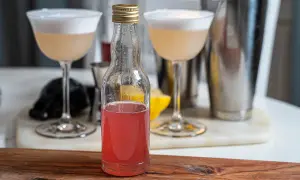 En flaska med rosa rabarbersirap står framför två drinkar med skum. En shaker och en citronklyfta syns i bakgrunden.