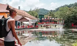 Mann mit Rucksack, der die bunte Malaysia Kleidung und die tropische Landschaft genießt. Ein See mit Fontäne und Brücke.