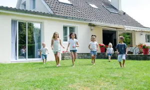 Dzieci biegają po trawniku przed domem z jasnym tynkiem zewnętrznym. Różne rodzaje tynków zewnętrznych mogą nadać budynkowi unikalny wygląd.