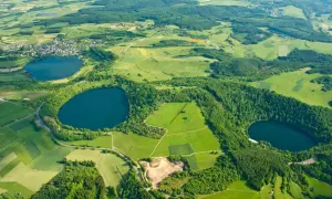 Luftaufnahme des **rundwegs um die drei Dauner Maare**. Grüne Felder, Wälder und drei dunkle Maare prägen die Landschaft.