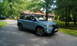 Jasnoniebieski Suzuki Vitara Hybrid zaparkowany na wybrukowanym podjeździe, otoczony zielenią i drewnianym budynkiem.