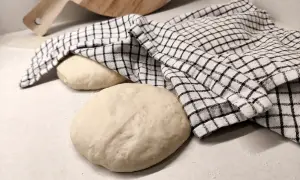 Två runda pizzadegar ligger på en bänk, en täckt av en rutig kökshandduk. Redo för bakning samma dag.