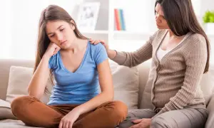 Smutna nastolatka siedzi na kanapie, a jej mama kładzie jej rękę na ramieniu, oferując wsparcie.