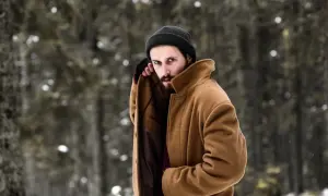 Mężczyzna w beżowym płaszczu i czarnej czapce, prezentujący stylowe zimowe stylizacje męskie na tle zaśnieżonego lasu.