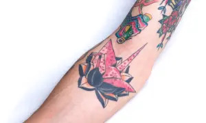 Kolorowe tatuaże na przedramieniu: różowy żuraw origami z kwiatami wiśni, róża i inne wzory w stylu yakuza.