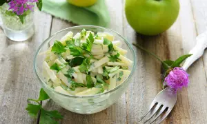 Frischer Kohlrabi Salat mit Petersilie in einer Glasschüssel, garniert mit einer lila Blume und grünen Äpfeln im Hintergrund.