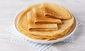 Pile de crêpes dorées, certaines pliées, sur une assiette blanche, posée sur une serviette à rayures bleues. Une délicieuse crêpe sans oeuf prête à être dégustée.
