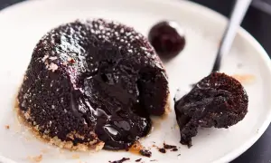 Un delizioso tortino al cioccolato con cuore fuso, cosparso di zucchero a velo, su un piatto bianco con una ciliegia e un cucchiaino.