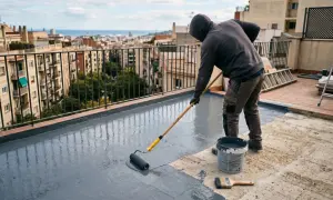 Hombre con capucha aplica pintura gris con rodillo para impermeabilizar pared exterior de terraza.