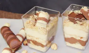 Deser Kinder Bueno w pucharkach z warstwami kremu, ciastek i czekolady, ozdobiony kawałkami batonika i orzechami.