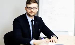 Mężczyzna w garniturze i okularach siedzi przy biurku, pisząc coś na kartce. Obok stoi filiżanka kawy.