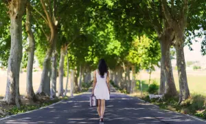 Vrouw in witte jurk loopt alleen over een laan met bomen, met een koffer in haar hand. Ze loopt weg, alsof ze weet dat ik val op mannen die mij afwijzen.
