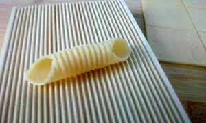 Frische Pasta selbst machen ohne Maschine: Ein geriffeltes Werkzeug formt Teig zu Röhren. Daneben liegen Teigplatten.